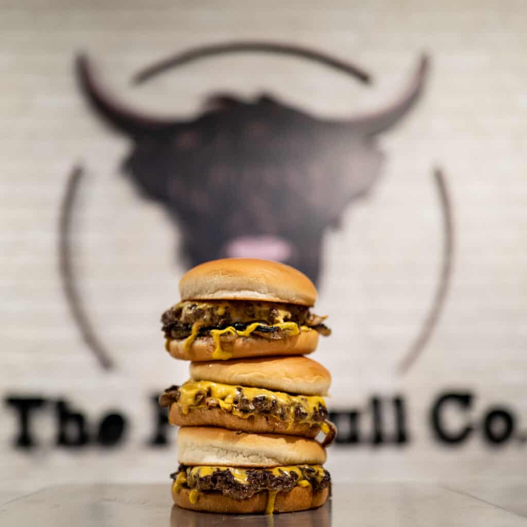 A tower of The Fat Bull Co., burgers​, stacked in front of the trademark Fat Bull logo, the highland cow.