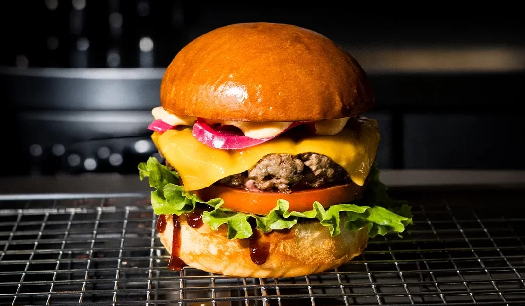 A juicy Fat Bull Co. burger with a hand-pressed beef patty, melted cheese, pickled red onion, lettuce and tomato on a toasted brioche bun, finished with a drizzle of sauce.